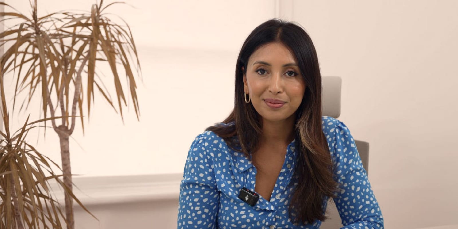 Dark-haired clinician in a patterned blue top talking to camera in a bright room beside a potted plant