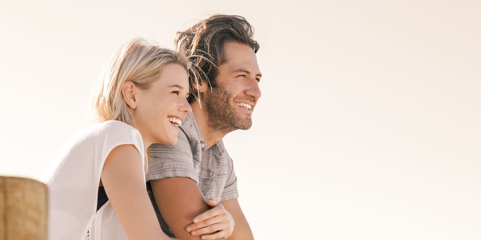 A couple leaning on a wooden railing outdoors in soft sunlight, smiling and looking into the distance